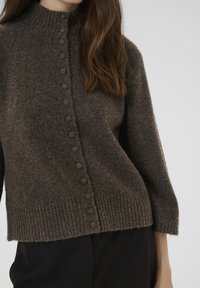 Egy nő sötétbarna gyapjú pulóvert visel, amelynek elülső részén kerek, anyaggal borított gombok találhatók, valamint bordázott mandzsetták és szegély.