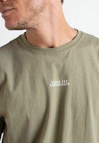 Oliven grønn bomulls t-skjorte med rund hals og hvit trykt tekst "CLEAN CUT COPENHAGEN" på brystet. Myk tekstur, avslappet design.