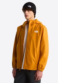 Veste imperméable jaune avec capuche et poignets élastiques, arborant un logo sur la poitrine et une fermeture éclair à l'avant. Portée avec un bonnet noir.