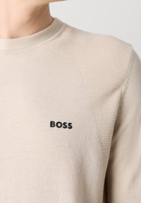 Pull en maille beige avec des panneaux côtelés aux épaules et un logo noir « BOSS » sur la poitrine gauche, porté par une personne légèrement tournée vers la gauche.