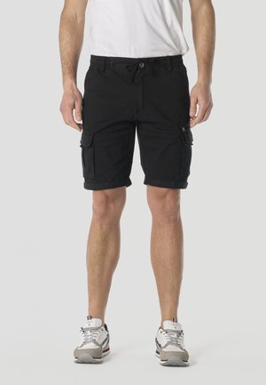 Adulto con pantalones cortos cargo negros con bolsillos con solapa y zapatillas blancas, de pie contra un fondo liso y claro.