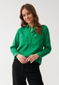 Cămașă verde cu nasturi, realizată dintr-un material neted, având două buzunare frontale, guler și mâneci lungi. Modelul o poartă cu pantaloni negri.
