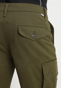 Pantalon cargo vert olive avec poches à rabat boutonnées à l'arrière et sur les côtés, porté avec un top noir.