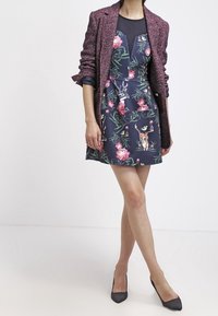 Merellinen kukkamekko, jossa on vaaleanpunaisia ruusuja ja peuroja, yhdistettynä tekstuuriseen burgundinpunaisiin bleisereihin. Mustat kärkitossut täydentävät asua.