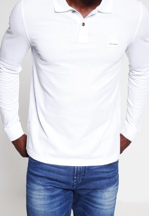 Bílá polokošile s dlouhým rukávem, se zapínáním na knoflíky a malou čtvercovou nášivkou s logem na hrudi, kombinovaná s modrými džínami.