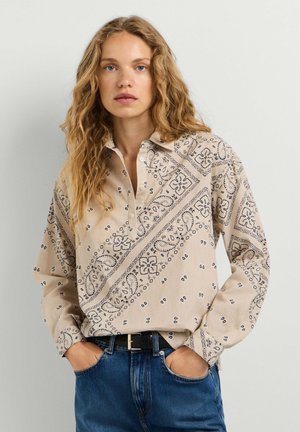 Femme aux longs cheveux bouclés portant une chemise beige à motifs boutonnée rentrée dans un jean bleu, les mains dans les poches.