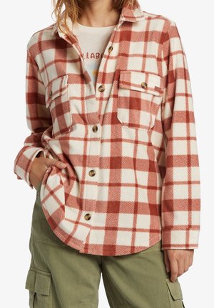 Kobieta w koszuli w kratę w kolorze rdza i białym, nosząca pod nią koszulkę z nadrukiem, w połączeniu z oliwkowozielonymi spodniami typu cargo, z dłonią w kieszeni.