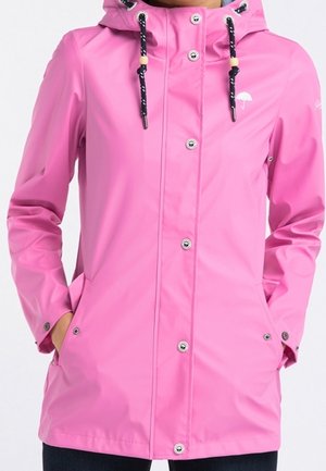 Veste imperméable rose avec capuche, cordons noirs, boutons-pression argentés et poches, portée avec les mains dans les poches.
