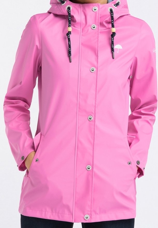Veste imperméable rose avec capuche, cordons noirs, boutons-pression argentés et poches, portée avec les mains dans les poches.