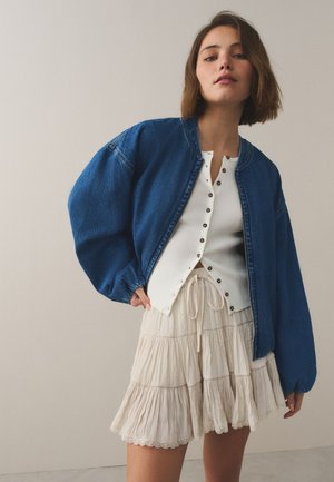 Jonge vrouw met een blauwe spijkerjas, wit geribd topje met knopen en een crèmekleurige gelaagde rok, staand tegen een effen achtergrond.