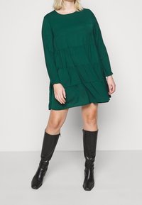 Abito verde a maniche lunghe con design a strati, scollatura rotonda e tessuto morbido, abbinato a stivali neri fino al ginocchio con punta quadrata.