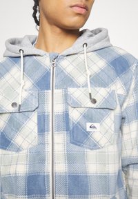 Personne portant une veste zippée à carreaux bleu et blanc avec des poches à rabat et une capuche grise avec des cordons blancs.