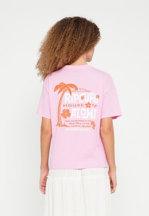 Femme aux cheveux bouclés portant un t-shirt rose avec un texte et des motifs hawaïens orange, associée à une jupe blanche, vue de dos.