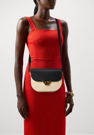 Kobieta ubrana w czerwoną sukienkę bez rękawów, z dwukolorową, czarno-beżową torebką na ramię oraz złotą bransoletką na prawej ręce.