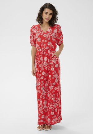 Femme aux cheveux bouclés portant une robe longue rouge à fleurs avec des manches bouffantes et des sandales, debout avec une main dans la poche.