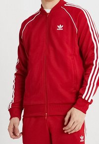 Punainen vetoketjullinen urheilutakki, joka on valmistettu sileästä kankaasta. Takissa on valkoiset raidat hihoissa ja brodeerattu logo rinnassa.