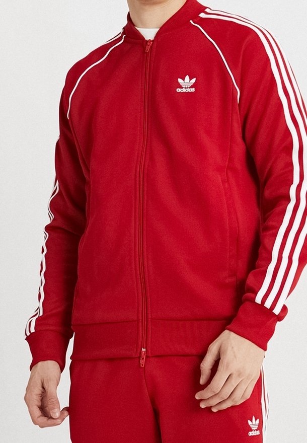 Punainen vetoketjullinen urheilutakki, joka on valmistettu sileästä kankaasta. Takissa on valkoiset raidat hihoissa ja brodeerattu logo rinnassa.