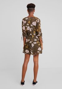 Robe florale à manches longues, en tissu vert olive, motif floral rose et jaune, liens ajustables aux manches, courte. Portée avec des baskets noires.