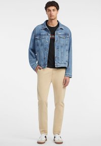 Hellblaue Jeansjacke mit zwei Brusttaschen, getragen über einem schwarzen T-Shirt, kombiniert mit beigen Chinos und weißen Sneakern mit schwarzen Akzenten.