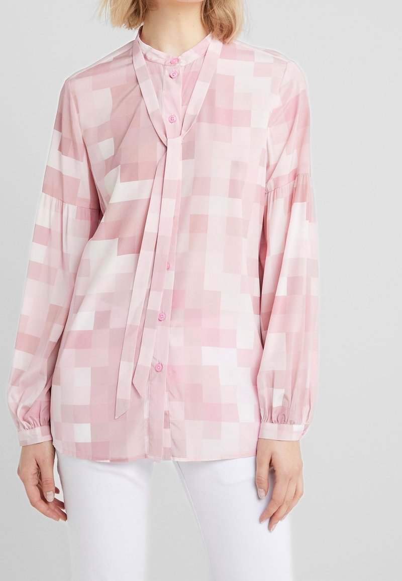 Femme portant un chemisier à motif géométrique rose clair et blanc avec manches longues et détail lavallière, associé à un pantalon blanc.