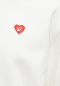 White sweatshirt with a round red fabric patch featuring "HUGO" in white text inside a heart shape, smooth texture, casual design.