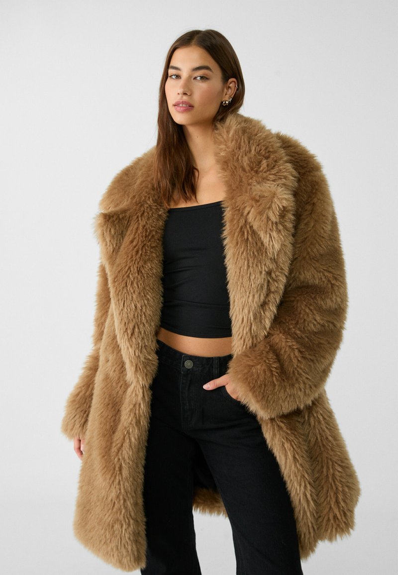 Cappotto in pelliccia ecologica marrone con ampio colletto, maniche lunghe e texture morbida. Indossato su una maglietta nera e abbinato a pantaloni neri.