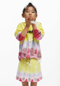 Felpa tie-dye gialla e rosa con testo, abbinata a una gonna tie-dye coordinata; tessuto morbido, vestibilità ampia e nappine decorative.