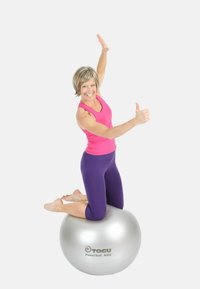 Srebrna fitness lopta označena "TOGU Powerball ABS", s ženom u ružičastom topu i ljubičastim tajicama, koja kleči i podiže ruku.
