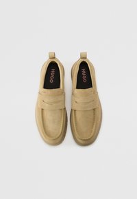 Loafers din suede bej cu o curea lată peste partea superioară, vârf rotund, branț moale și tab de prindere la spate; numele mărcii imprimat în interior.