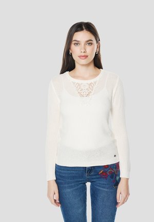 Suéter de punto blanco de manga larga, con un panel de encaje en el escote. Combinado con jeans azules decorados con bordados florales.