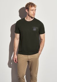 Musta vihreä pyöreäkauluksinen t-paita, joka on valmistettu puuvillasta ja jossa on pieni painettu etikettisuunnittelu vasemmassa yläkulmassa. Yhdistetty vaaleanbeigeihin housuihin.