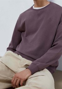 Bluza w stonowanym fioletowym kolorze z bawełny. Klasyczny okrągły dekolt, ściągacze przy mankietach i u dołu, z delikatnym tonalnym logo na piersi. W połączeniu z beżowymi spodniami.