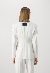 Biała, dopasowana marynarka z usztywnionymi ramionami i dopasowanym tyłem. Na obszarze szyi znajduje się czarna metka. Gładka tkanina o czystych liniach.