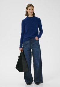 Modrý pletený sveter s textúrovaným povrchom, skombinovaný so širokými tmavými džínsami. Modelka drží čiernu kabelku. Jednoduchý a moderný dizajn.