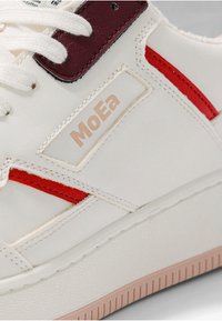 Witte sneaker met rode accenten, glad synthetisch bovenwerk, verhoogd logo "MoEa," en een getextureerde rubberen zool met een lichtbruine onderkant.