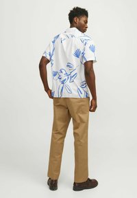 Camisa de manga corta de algodón blanco con estampado floral azul; combinada con pantalones marrones claros y zapatos oscuros; modelo visto por detrás.