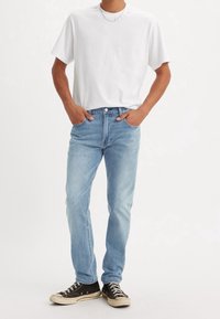 Homme portant un t-shirt blanc uni, un jean bleu clair, des baskets noires basses, debout les mains dans les poches sur fond blanc.