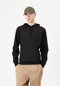 Hanorac negru cu glugă, cu un design elegant, având șnur și un accent mic cu logo galben. Purtați cu pantaloni bej.