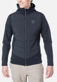 Musta huppari, jossa on vetoketjukiinnitys, edessä tikatut paneelit, sileä tekstuuri ja hillitty logotunnus rinnassa.