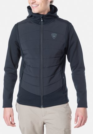 Veste à capuche noire avec fermeture éclair, dotée de panneaux matelassés sur le devant, d'une texture lisse et d'un détail de logo discret sur la poitrine.