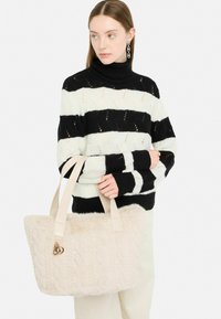 Sac fourre-tout en fausse fourrure beige avec des poignées en cuir lisse et un design en mailles texturées, tenu par une personne portant un pull rayé noir et blanc.