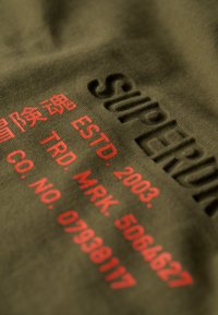 Tela verde oliva con el logotipo emblema "SUPERDRY" en relieve y texto impreso en rojo, que incluye "ESTD. 2003" e información de la empresa, creando un detalle texturizado.