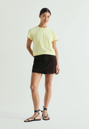 Mujer joven con una camiseta amarilla pálida con relieve de calavera, minifalda negra con botones y sandalias planas negras con cordones, de pie frente a un fondo sencillo.