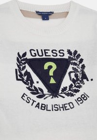 Fehér pulóver, kerek nyakú, fekete "GUESS" logóval, zöld kérdőjellel díszített háromszög mintával és babérlevelekkel kiemelve.