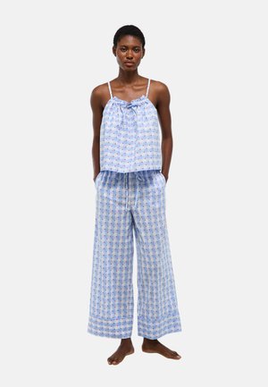 Femme portant un pyjama bleu clair à motif à carreaux rouges, débardeur sans manches avec cordon au col et pantalon à jambes larges, debout pieds nus.