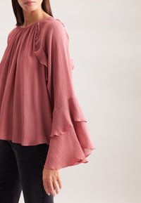 Blusa rosa sgualcita con un motivo testurizzato, vestibilità ampia e maniche larghe, abbinata a jeans scuri.
