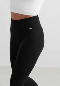 Gros plan d’une personne portant des leggings noirs côtelés taille haute et un top court blanc sur un fond clair uni.