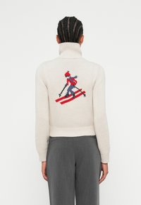 Pull léger beige avec un col montant. Il présente un motif de ski en rouge, bleu et noir au dos. Manches et ourlet côtelés.