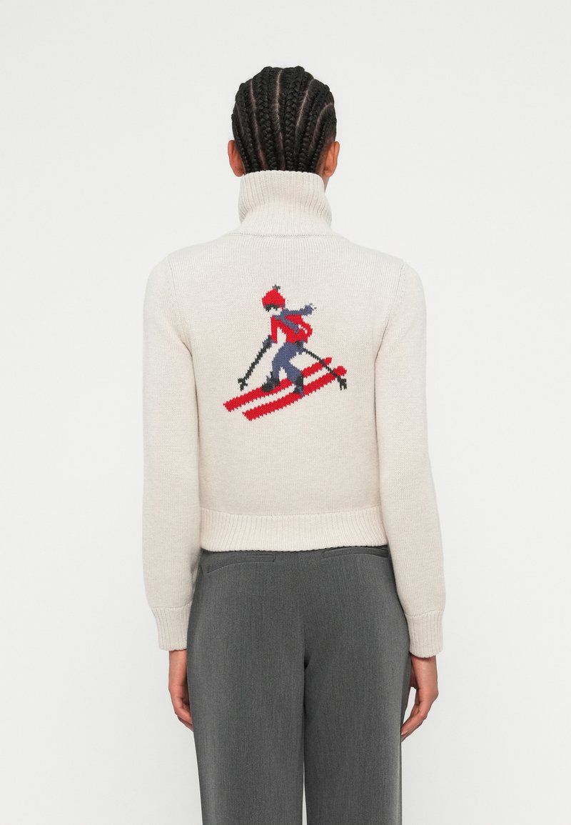 Pull léger beige avec un col montant. Il présente un motif de ski en rouge, bleu et noir au dos. Manches et ourlet côtelés.