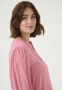 Blouse rose texturée avec des manches bouffantes, présentant un décolleté en V et des motifs à pois sur l'ensemble. Tissu lisse avec un éclat subtil.